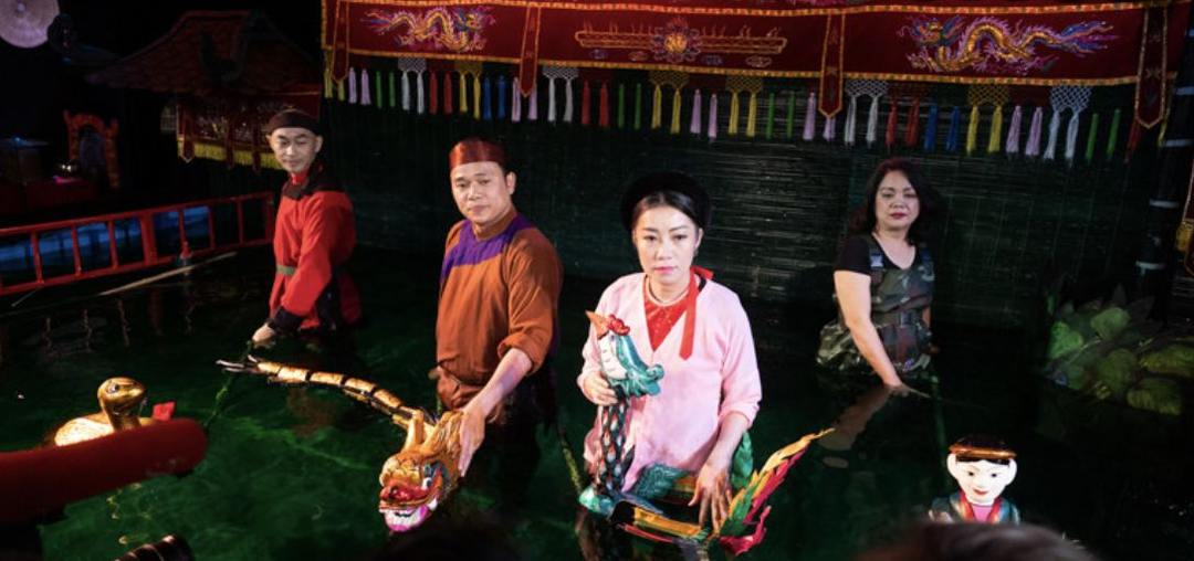 Bốn nghệ nhân đứng ngập người trong nước phía sau sân khấu, tay cầm các con rối rồng và rắn